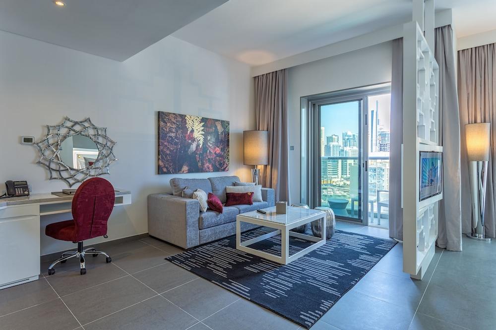 Hotel The First Collection Dubai Marina Esterno foto The photo shows a modern living space designed with a contemporary aesthetic. It features a comfortable gray sofa with colorful cushions, a stylish glass coffee table, and a dark patterned rug on the floor. There is a small desk with a red chair posi